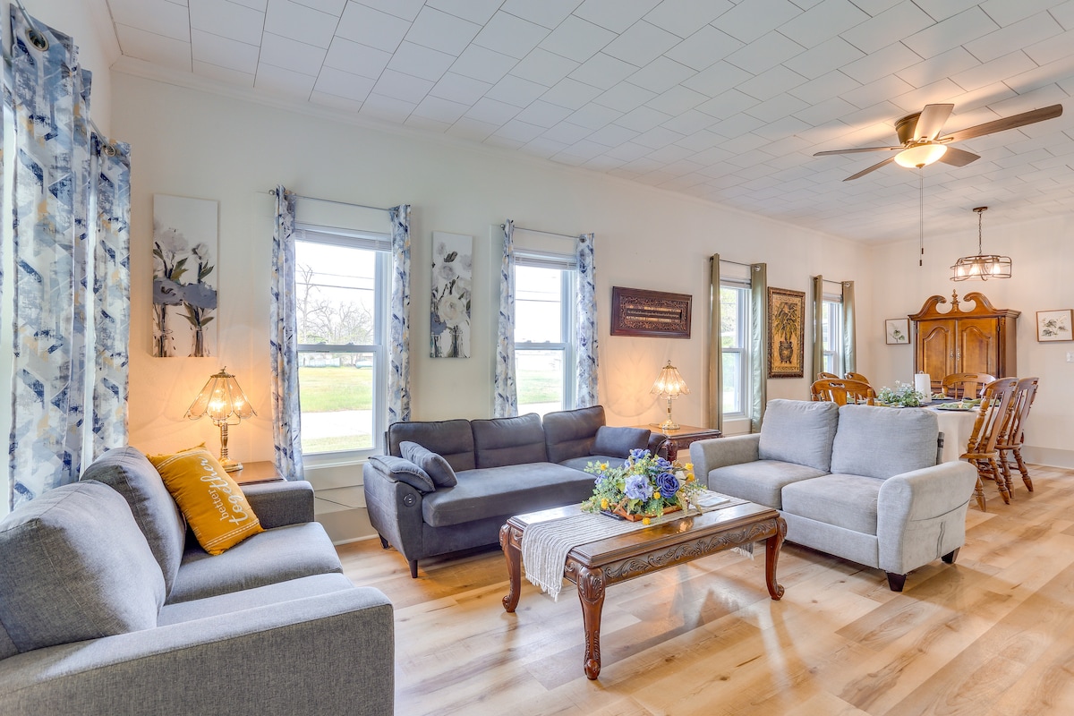 The living area features multiple seating options, including two grey couches and a decorative coffee table. Natural light enters through several windows adorned with floral curtains. A dining space is visible in the background, complete with wooden chairs and a hutch, enhancing the communal atmosphere.