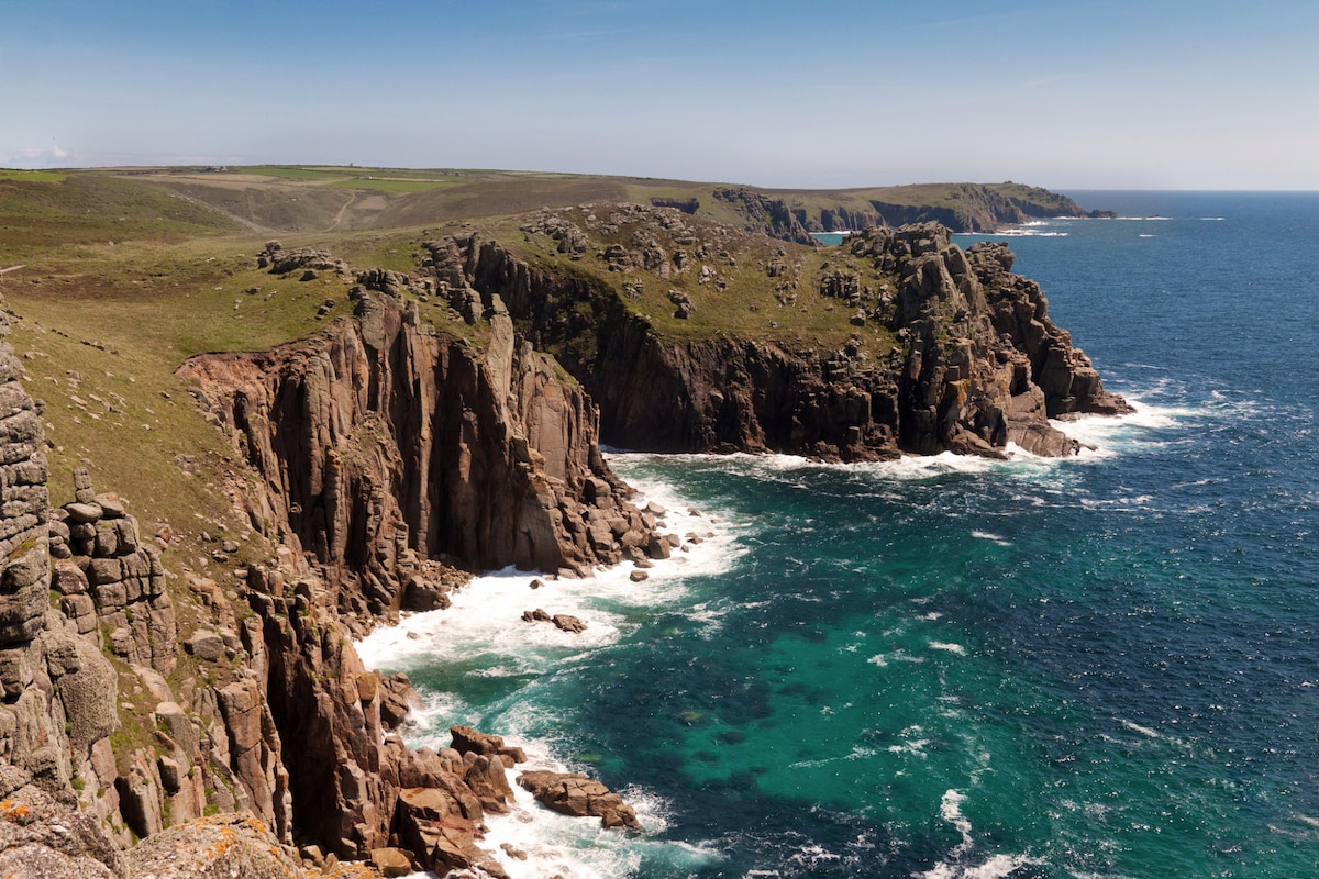 A rugged coastline is depicted, featuring steep cliffs that rise sharply from the turquoise waters below. Waves crash against the rocky shore, creating white foam. The landscape is enhanced by green hills in the background, contrasting with the deep blue sea under a clear sky.