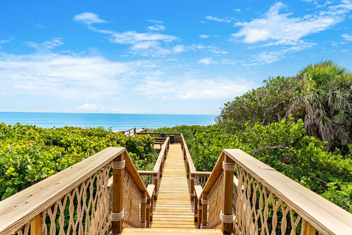 A wooden walkway leads through lush greenery towards the sandy beach, with the glistening ocean visible in the distance under a clear blue sky. The pathway is framed by vibrant plants, creating a serene connection to the natural surroundings.