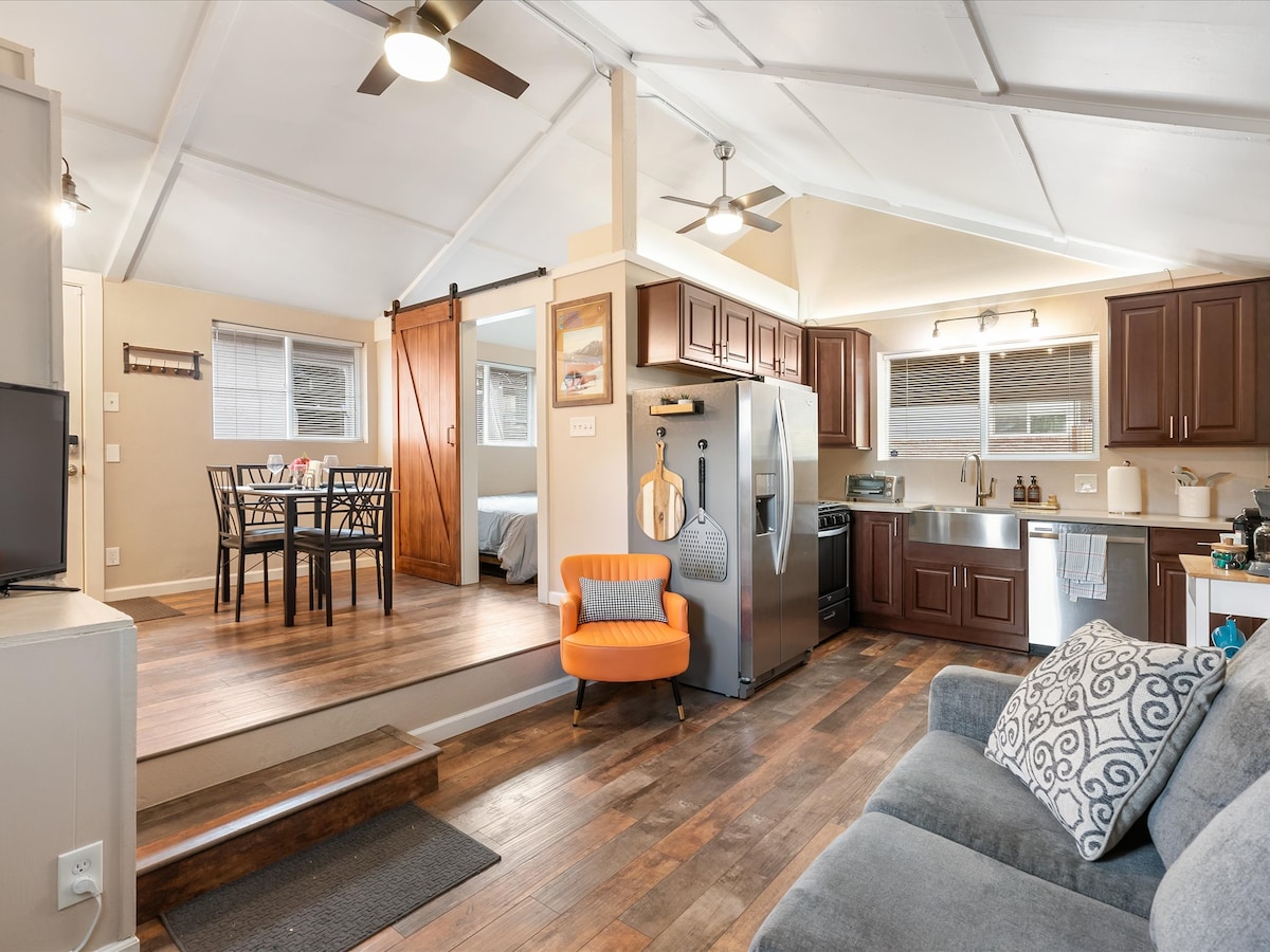 An open living area showcases a comfortable seating arrangement and a modern kitchen featuring stainless steel appliances and wooden cabinetry. A dining table for four is positioned nearby, with natural light illuminating the space through multiple windows.