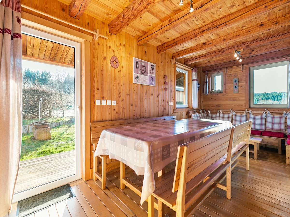 A spacious dining area is showcased, featuring a large table surrounded by wooden benches. Natural light floods in through a glass door leading to the garden, while wooden beams and paneling create a warm atmosphere. Art pieces adorn the walls, enhancing the cozy setting.