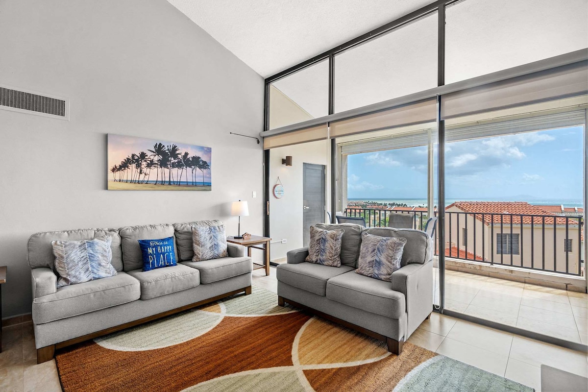 A spacious living area features two comfortable gray sofas facing a large glass door that opens to a private balcony with ocean views. A multi-colored area rug adds warmth, and a palm tree artwork enhances the space's relaxing ambiance.