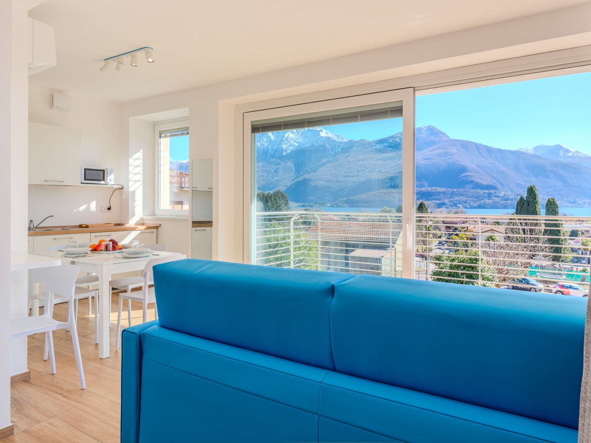 A bright living area showcases a comfortable blue sofa and a dining table with white chairs. A panoramic window offers a stunning view of the mountains and the lake, while natural light floods the space, enhancing the modern design.