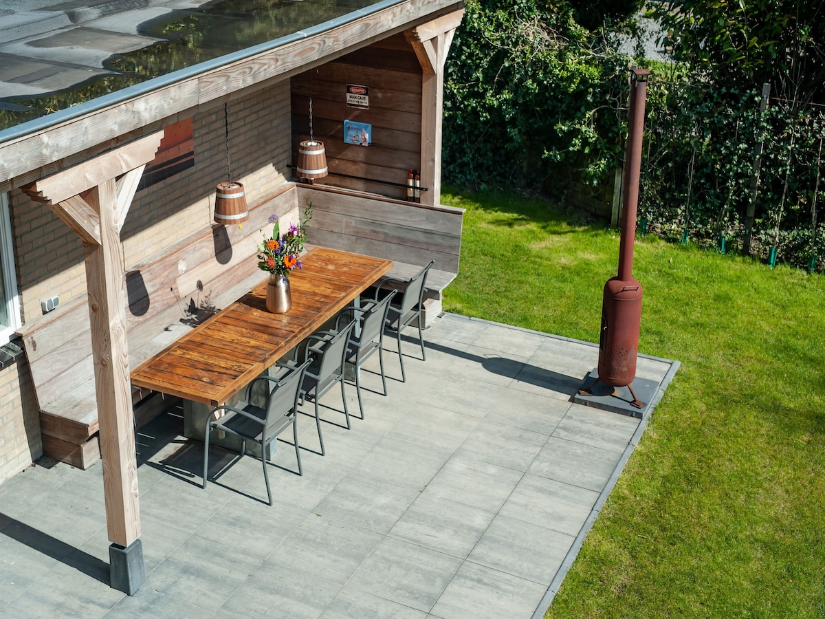 An outdoor terrace area is highlighted, featuring a large wooden dining table surrounded by several chairs. A rustic fireplace is positioned nearby, and pots of bright flowers add a touch of color. The green grass and surrounding greenery provide a sense of privacy.
