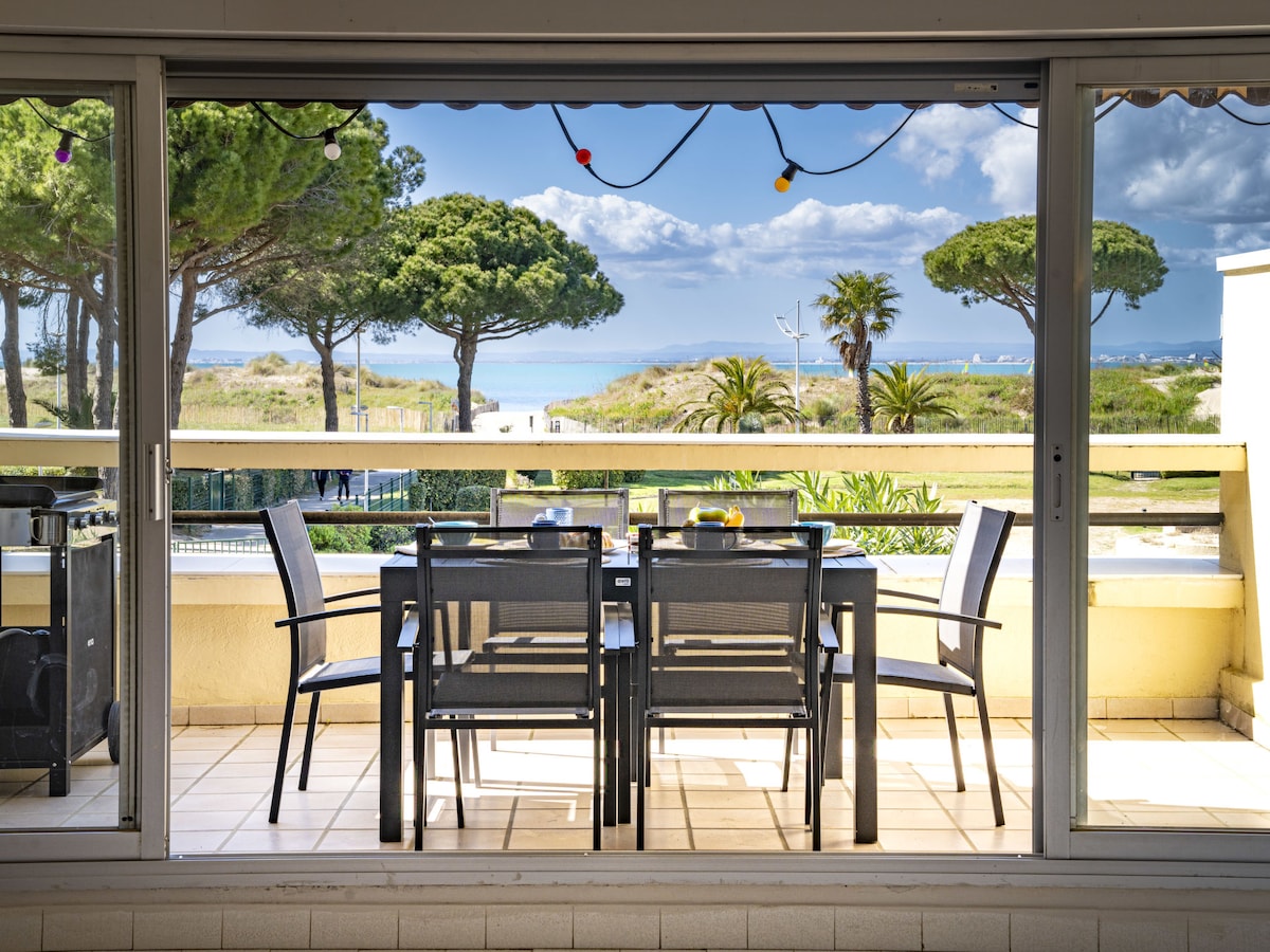 A terrace area is shown, furnished with a dining table and six chairs. Views of trees and the sea are visible beyond the railing, under a partly cloudy sky. An electric barbecue is positioned to the side, adding to the outdoor dining experience.