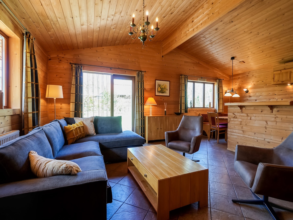 A spacious living and dining area features wooden walls and a high ceiling. Comfortable seating includes a large sectional sofa and two armchairs. A wooden coffee table occupies the center, while warm light illuminates the space from a chandelier and nearby lamps. Large windows provide natural light.