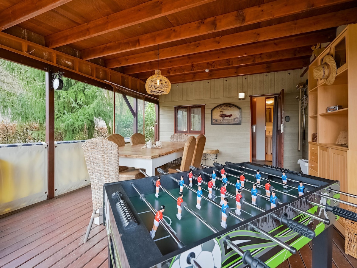 A covered terrace features a foosball table prominently in the foreground, with a dining table and wicker chairs arranged nearby. The space is illuminated by natural light, with wooden beams overhead and a doorway leading to additional areas of the chalet.