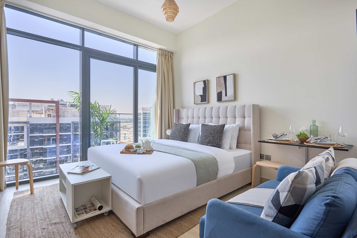 A bright studio apartment is featured, showcasing a spacious bed with soft linens and decorative pillows. Floor-to-ceiling windows invite natural light, providing a view of the cityscape. A cozy blue loveseat and a compact dining area are visible, enhancing the living space's inviting layout.