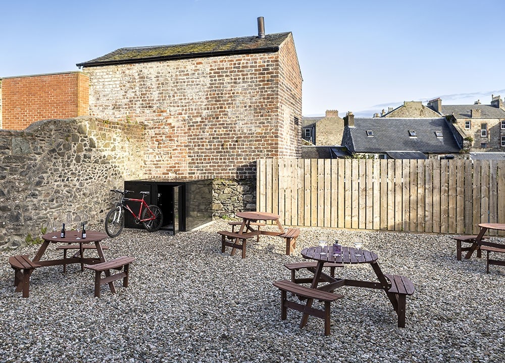 An enclosed walled garden area features several picnic benches arranged on gravel. A bike locker is visible against the brick wall, providing storage for bicycles. The surrounding fencing and buildings create a private atmosphere, ideal for outdoor relaxation.