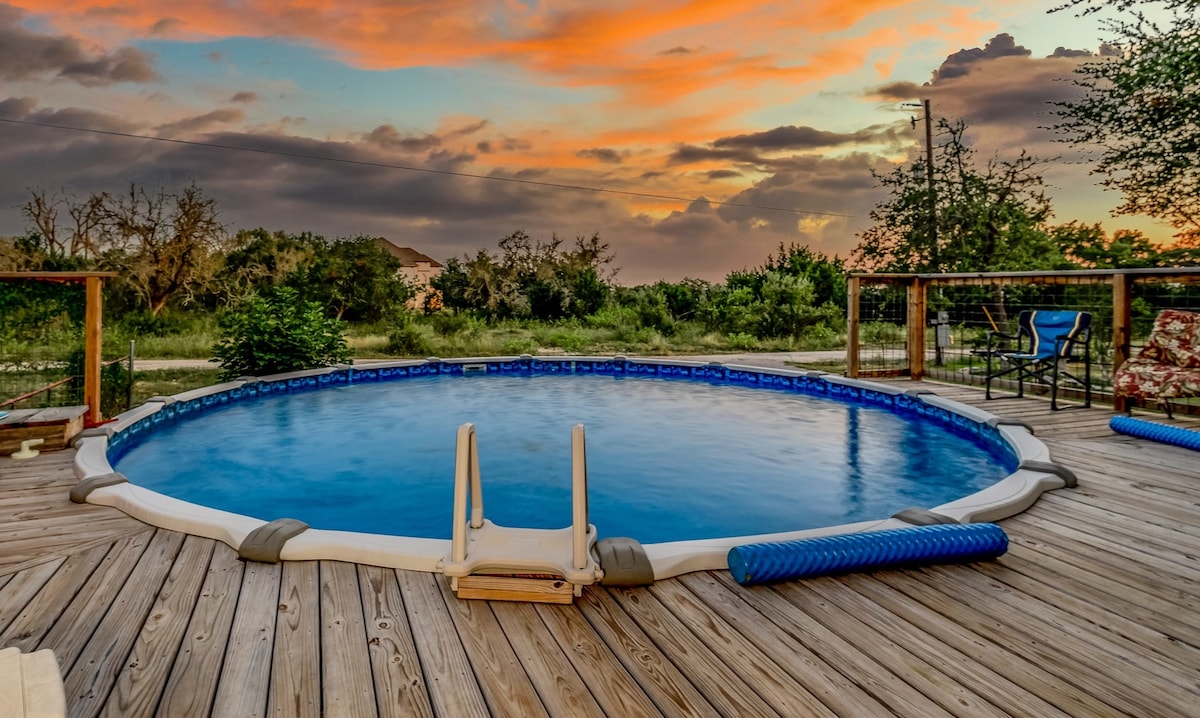 A circular swimming pool is set on a wooden deck, surrounded by lush greenery and a scenic landscape. Two lounge chairs and a seating area are visible, with an inviting sunset reflecting off the water's surface.