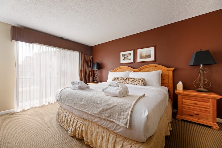 Удобна спална соба со топли тонови, мека постелнина и нежно осветлување покрај креветот.