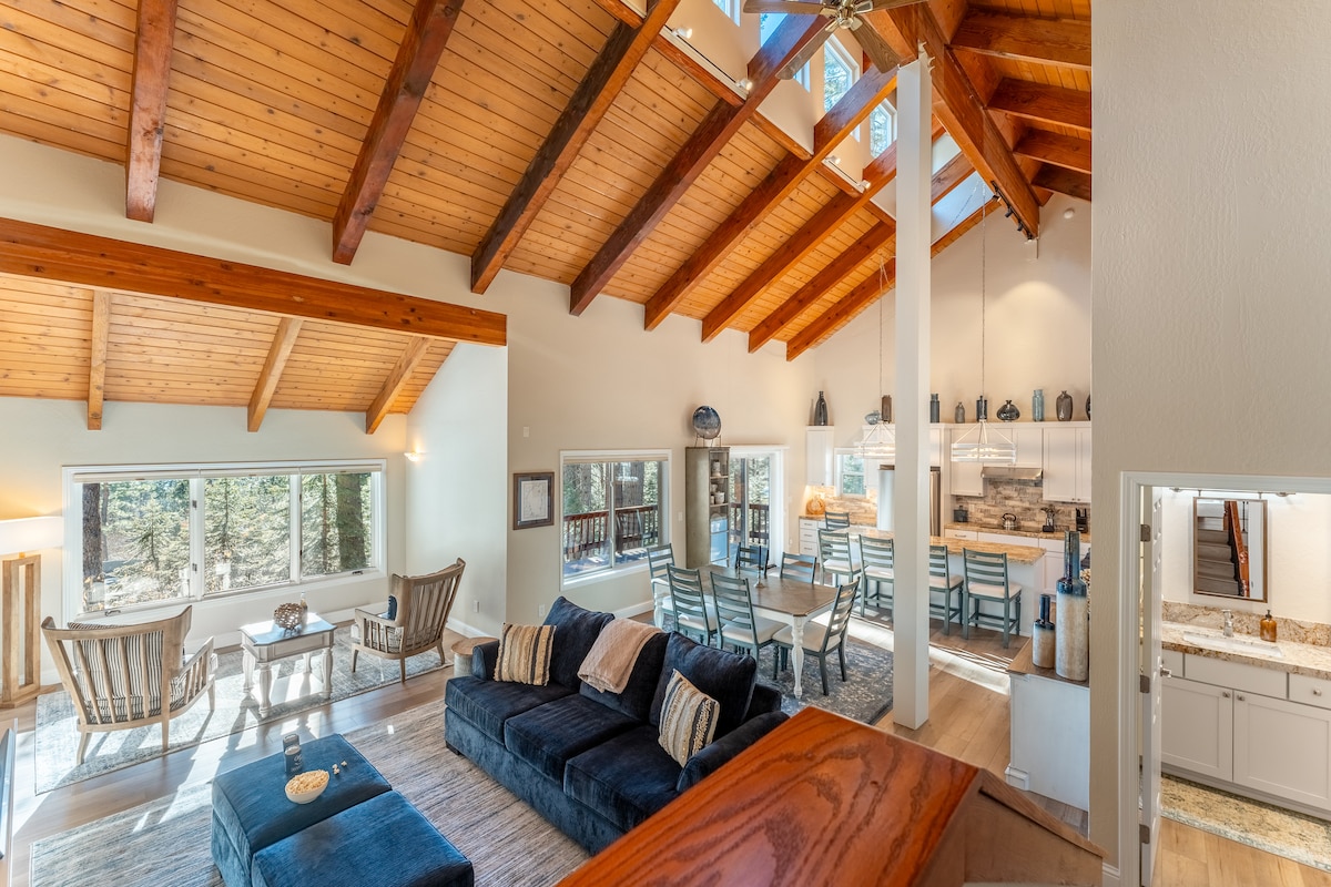 The expansive living area features high ceilings with exposed wooden beams and large windows that invite natural light. A spacious sectional sofa faces a coffee table, while a dining area with multiple chairs and a fully equipped kitchen is visible in the background.