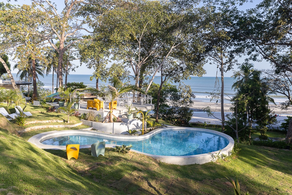 A tranquil pool area is surrounded by lush greenery and palm trees, offering views of the ocean in the background. Comfortable lounge chairs are positioned for relaxation, inviting guests to unwind while enjoying the natural beauty of the coastal landscape.