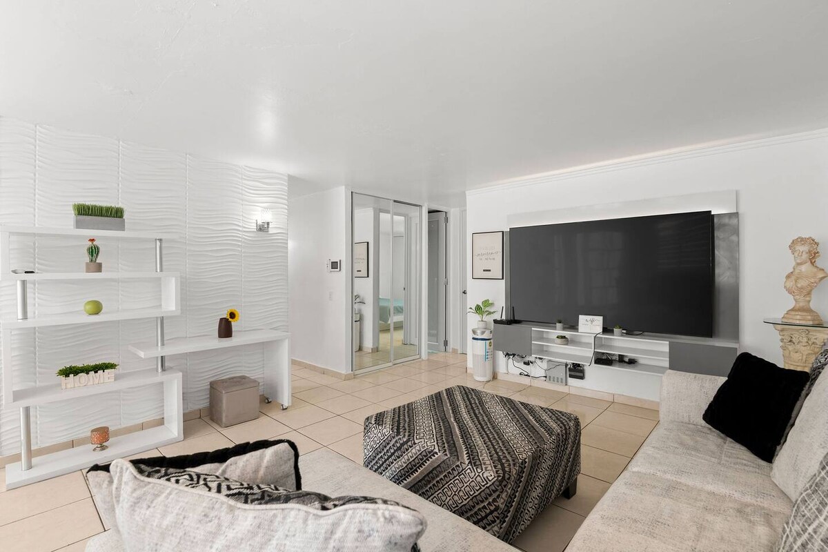 A spacious living area features a large wall-mounted television with a sleek media console. Comfortable seating arrangements include a textured ottoman and a cozy sofa. Light-colored tiles create a bright atmosphere, complemented by minimalist shelving displaying decorative items.