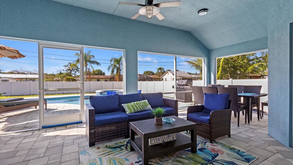 A screened-in back porch offers a spacious seating area with comfortable wicker furniture and a decorative rug. Natural light floods in through large windows, showcasing the private heated pool and fenced backyard beyond. A dining table is positioned nearby for outdoor meals.