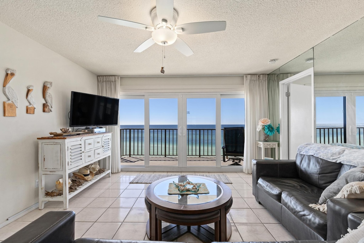 A spacious living area features large glass doors offering sweeping views of the Gulf. Comfortable seating includes dark leather couches, and a modern coffee table is placed in the center. Natural light fills the room, enhancing the coastal decor.