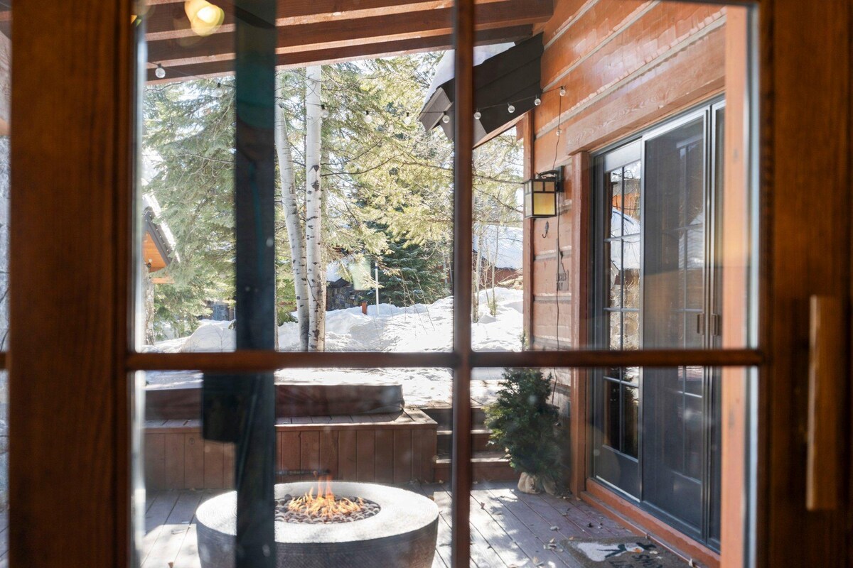 The view through the glass-paneled doors reveals a private patio area featuring a circular fire pit. Surrounding trees provide a natural backdrop, while snow blankets the ground, creating a serene winter atmosphere.