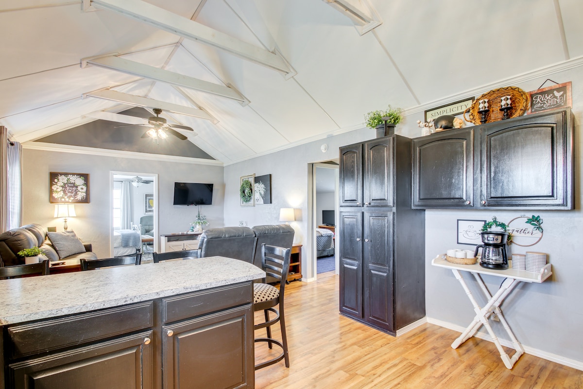 The open-concept living area features vaulted ceilings and a stylish kitchen with dark cabinetry. A breakfast bar provides seating and a view of the comfortable lounge, while large windows allow natural light to fill the space. A cozy atmosphere is created with light-colored walls and warm wood flooring.