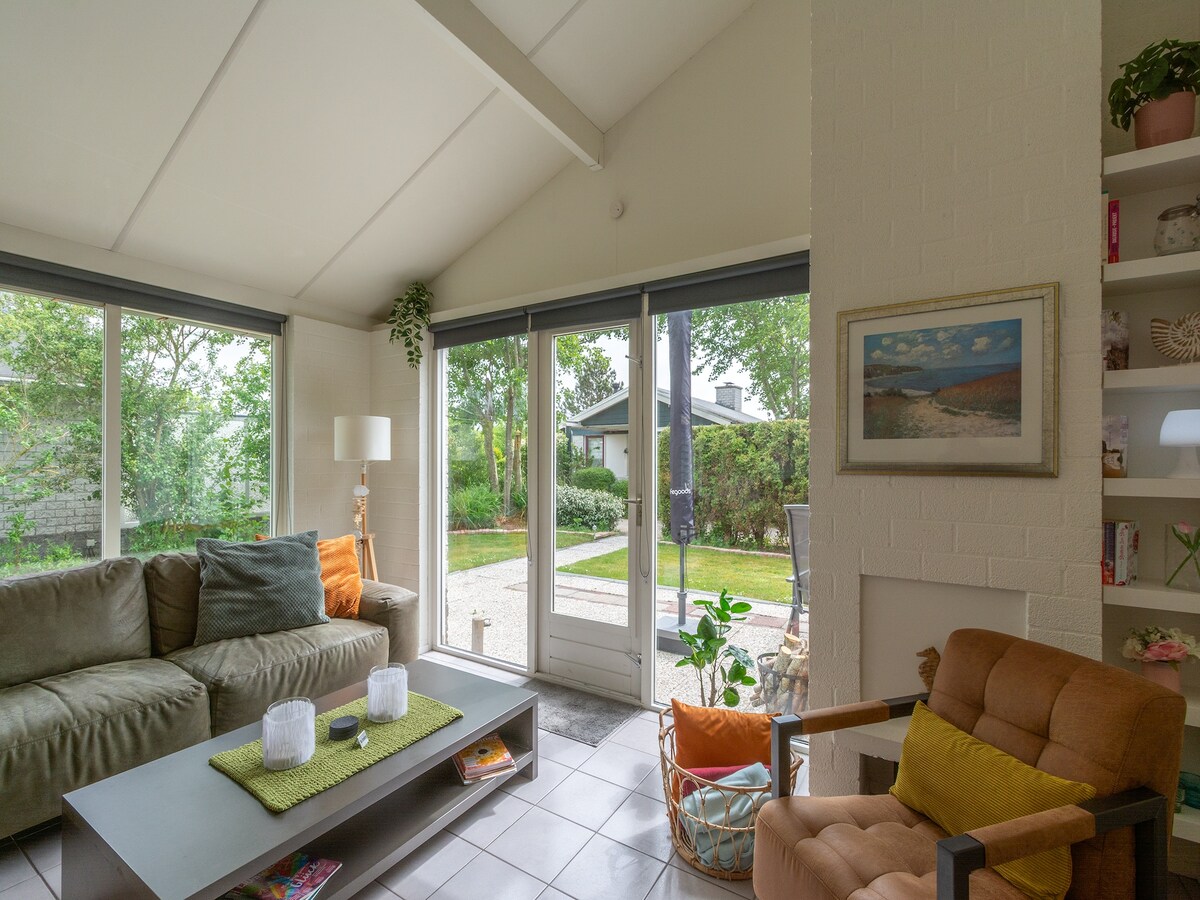A cozy living room features a sectional sofa and an armchair, creating a welcoming seating area. Large windows provide a view of the garden, and natural light fills the space. A coffee table sits in the center, with decorative items adding a personal touch.