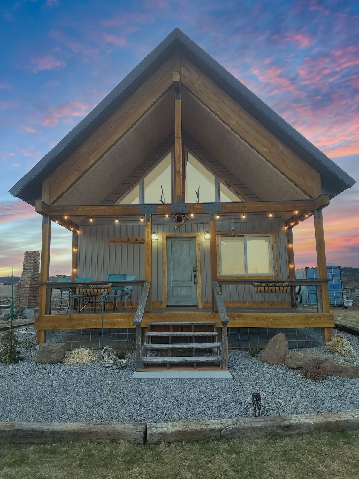 A charming cabin features a peaked roof and large angled windows, providing ample natural light. The wooden porch with steps is adorned with decorative lights. Surrounding gravel and rocks create a rustic landscape, while a colorful sunset adds warmth to the scene.