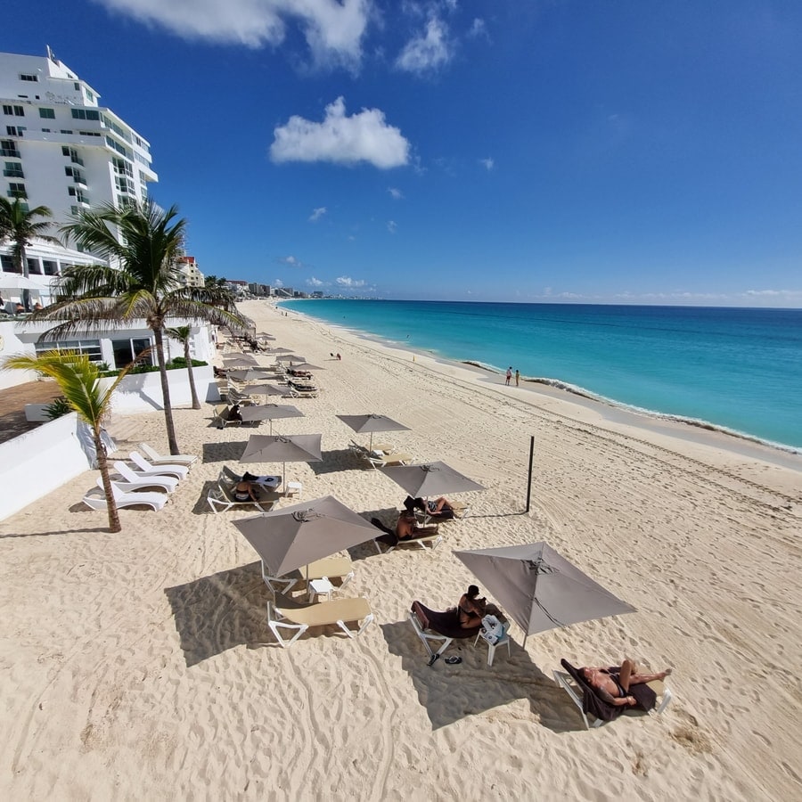 A serene beach scene captures numerous lounge chairs and umbrellas aligned along a pristine sandy shore. The turquoise ocean gently laps at the beach, with a clear blue sky overhead. A few beachgoers can be seen enjoying the sun and tranquility.