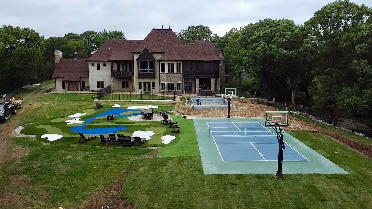 The expansive outdoor area presents a well-manicured lawn featuring a mini golf course with greens and bunkers. A basketball court with two hoops is visible, alongside a large patio area with seating that provides a view of the lodge. The lodge features a combination of stone and wood elements.