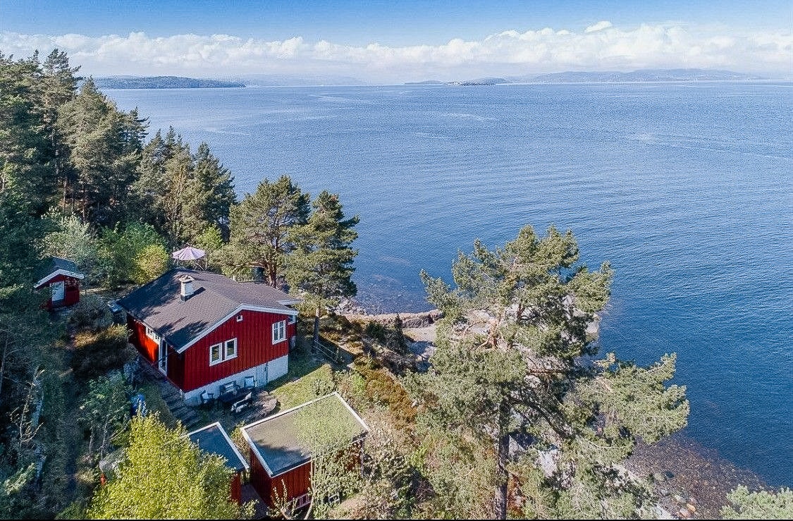 An aerial view of a charming red cabin nestled among trees, with the serene Oslofjord extending into the horizon. The clear waters reflect the sky, while the surrounding landscape includes rocky shorelines and greenery, creating a tranquil ambiance.