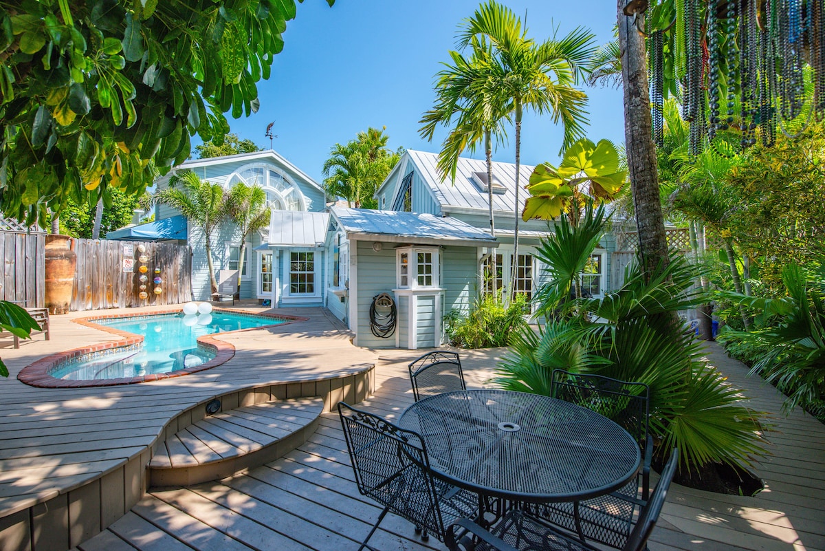 A private outdoor space is featured with a wooden deck surrounding a sparkling pool. Lush greenery, including palm trees and shrubs, enhances the serene environment. A circular dining table with black chairs is positioned nearby, offering a perfect setting for outdoor meals or relaxation.