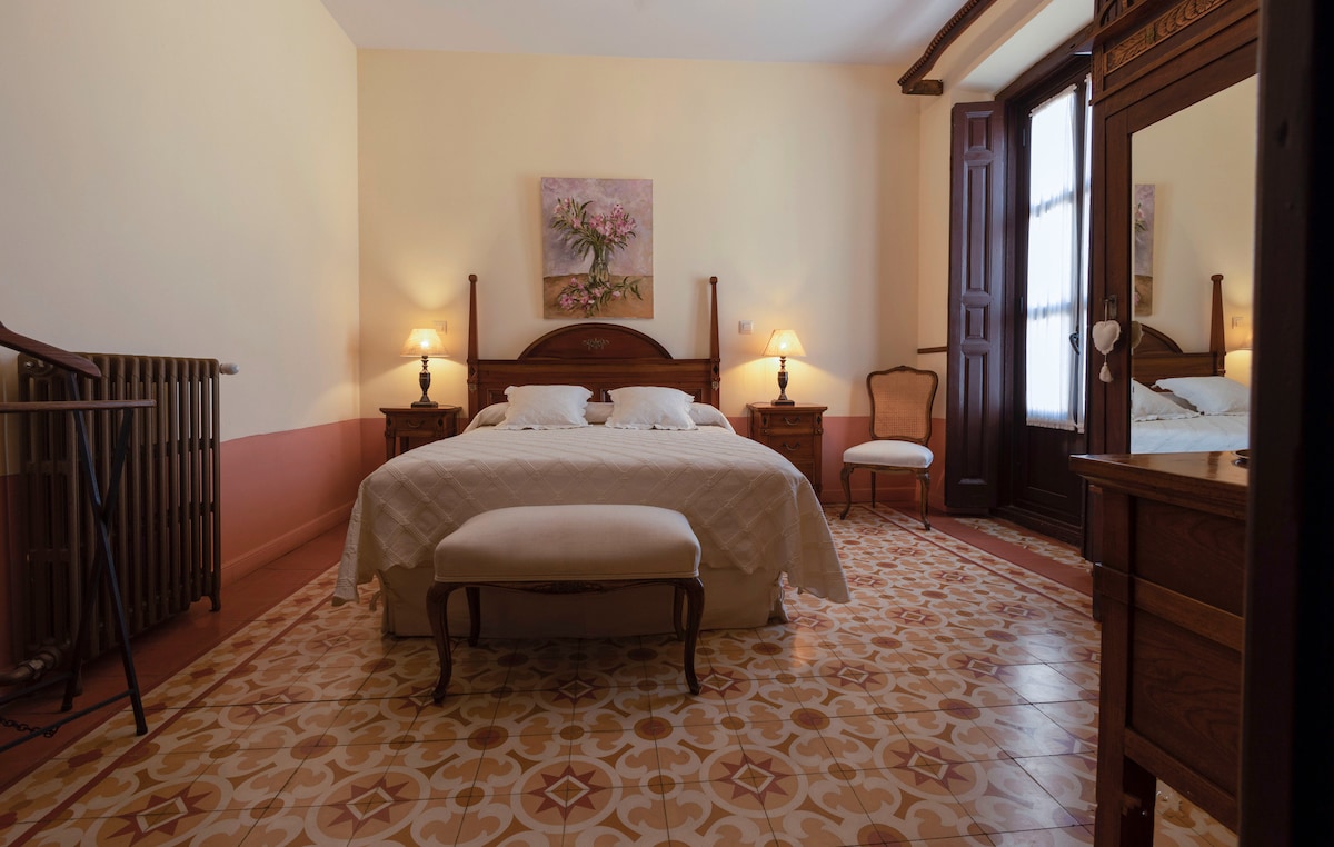 A spacious bedroom is featured with a large bed flanked by wooden nightstands and lamps. A decorative painting complements the warm yellow walls. A cozy seating area is provided, and tall windows allow natural light to enhance the ambiance.