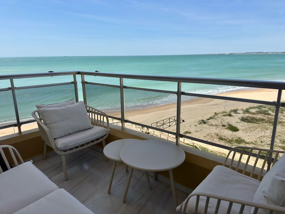 A serene terrace is presented, featuring modern white seating and a round table. Breathtaking views of the sandy beach and turquoise waters extend beyond the glass railing, offering a calming beachfront atmosphere that invites relaxation.