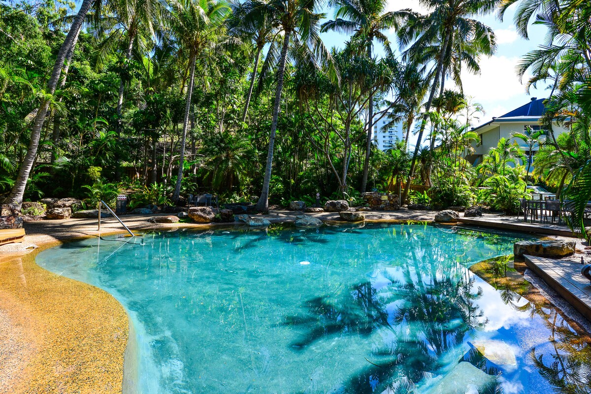 A refreshing swimming pool is surrounded by lush greenery and swaying palm trees, creating a serene atmosphere. Natural stone elements border the pool, reflecting the clear blue water and sunlight, inviting relaxation and leisure in a tropical setting.