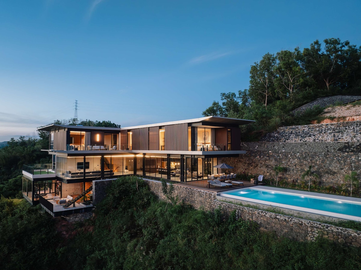 A modern villa is nestled on a hillside, showcasing a combination of glass and wood for a sleek appearance. An infinity pool reflects the sky, while the surrounding greenery enhances the tranquil setting. An expansive terrace offers an inviting space for relaxation and gatherings.