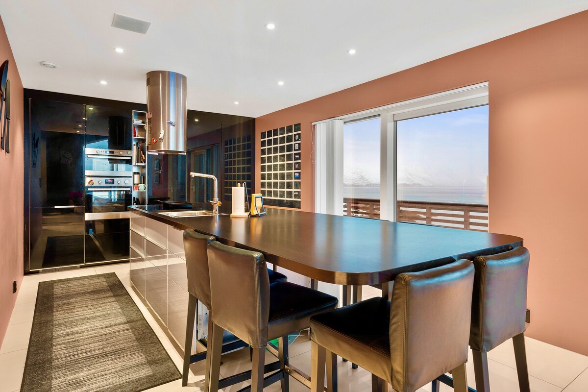 A modern kitchen is highlighted by a long dining table with dark leather chairs. Sleek cabinets and high-end appliances are visible along one wall, while large windows offer a clear view of the outside landscape. Soft lighting enhances the clean lines of the space.