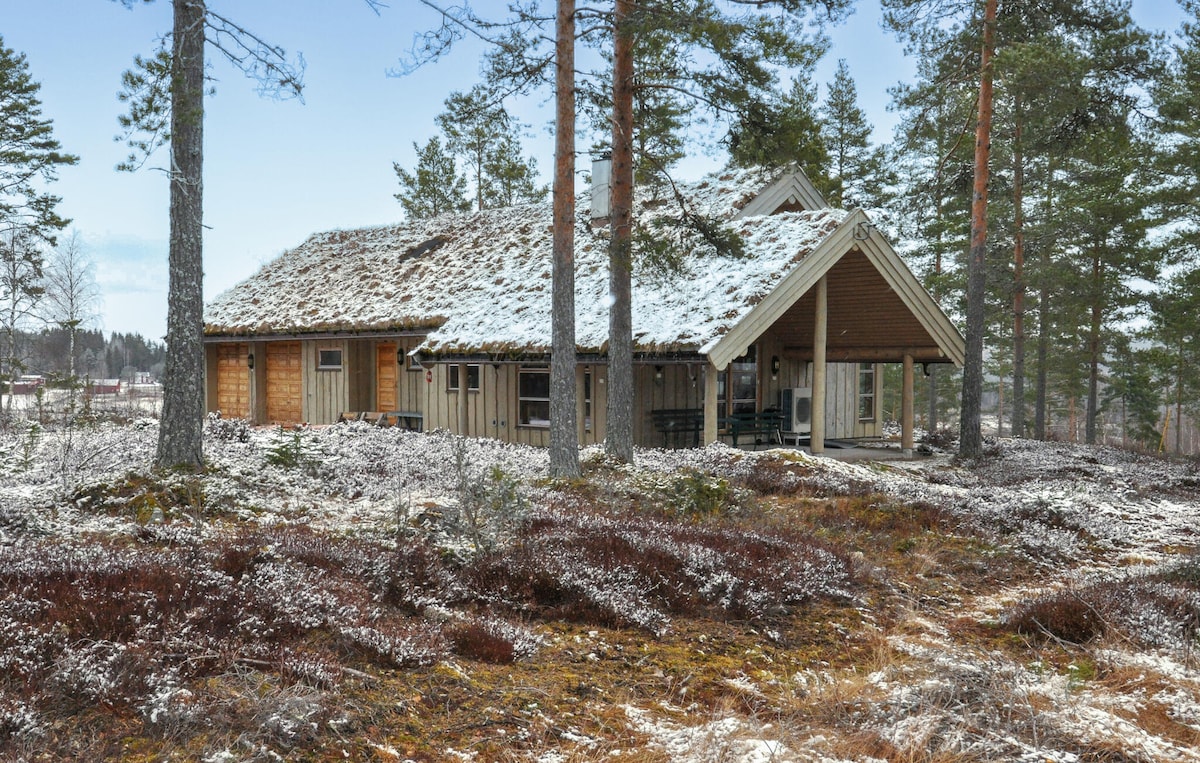 A charming holiday home is set amid a natural landscape, surrounded by tall trees and patches of snow. The rustic exterior, featuring wooden siding and a sloped roof, complements the serene forest environment. A covered porch extends outwards, inviting outdoor enjoyment.