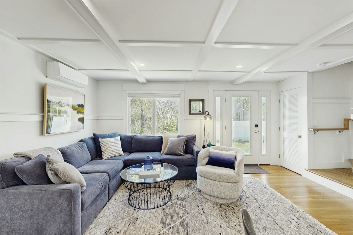 A cozy living area is presented with a large sectional sofa, complemented by a round glass coffee table. Natural light filters in through expansive windows, illuminating the neutral decor. A stylish armchair sits nearby, creating an inviting space for relaxation and comfort.