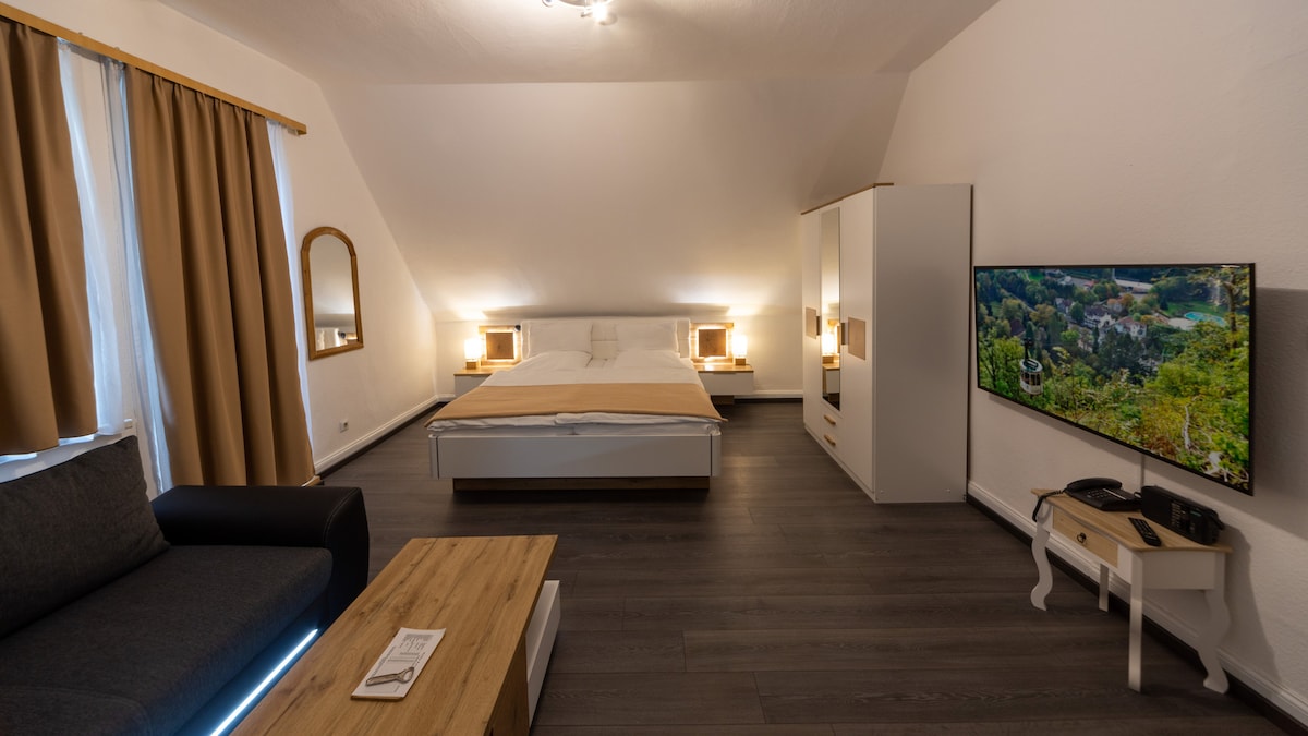 A spacious bedroom is furnished with a large bed flanked by two bedside lamps. A modern flat-screen TV is mounted on the wall opposite a cozy seating area, which includes a dark sofa and a wooden coffee table. Natural light filters through beige curtains.