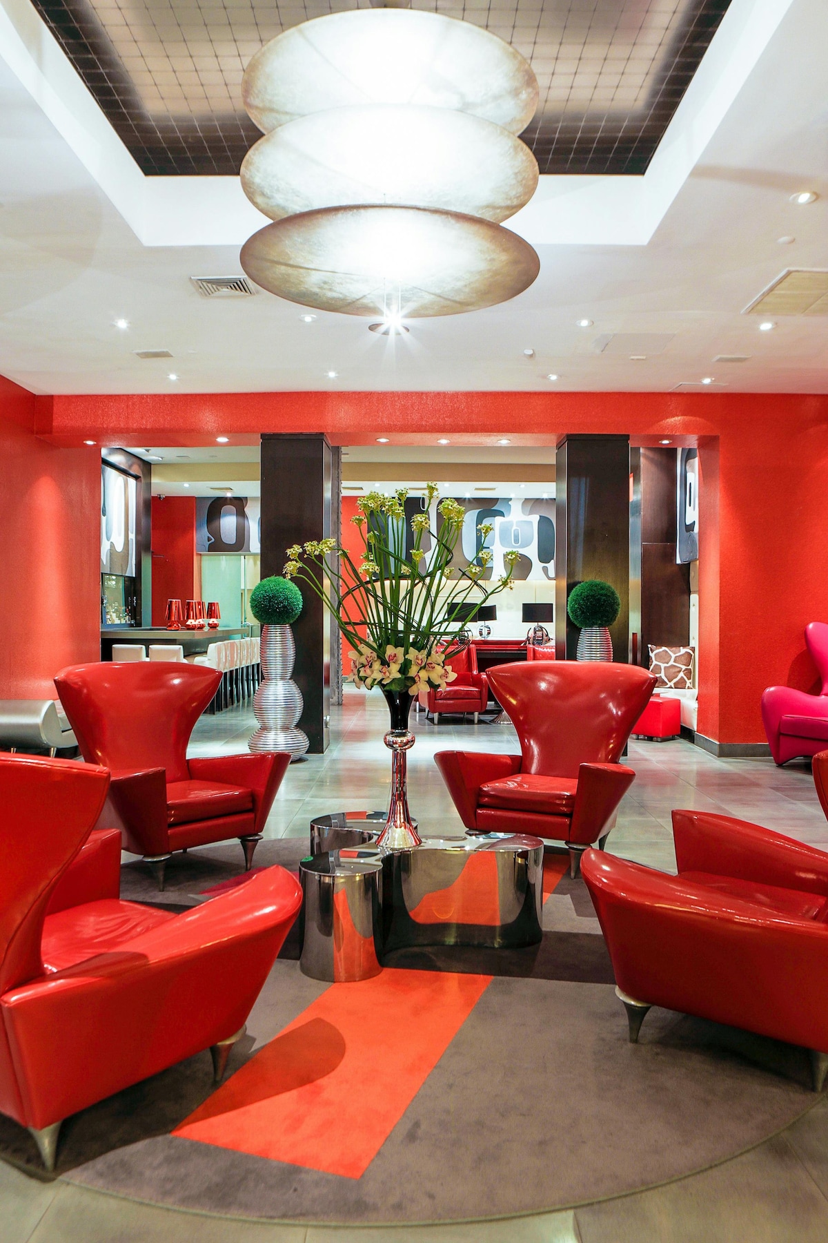 A vibrant lobby area features red accent walls and modern furnishings. Two bold red chairs are positioned around a circular table, complemented by a decorative floral arrangement. Bright overhead lighting highlights the sleek design, while soft carpeting adds comfort underfoot.