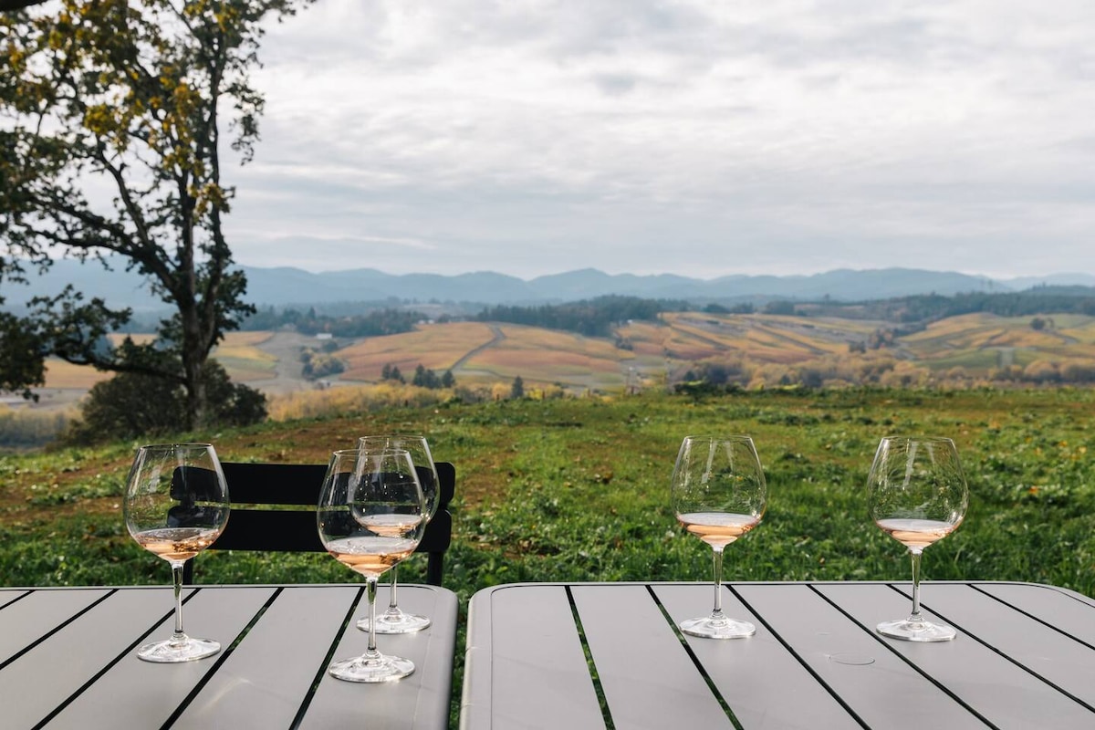 A scenic view of rolling hills showcases the lush countryside, with four stemmed glasses of rosé arranged on a white table. The landscape features patches of green and gold, contrasted against a cloudy sky that casts a soft light over the vibrant terrain.