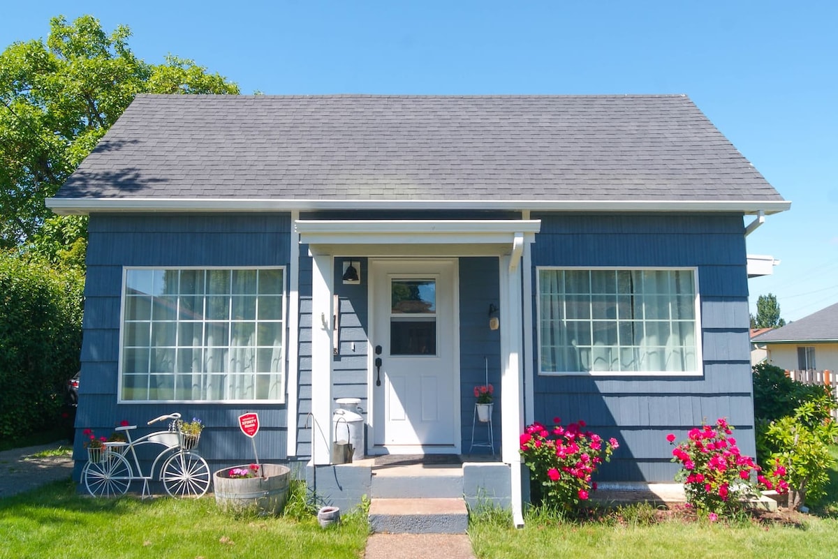 A charming blue cottage is viewed from the front, showcasing a well-maintained lawn dotted with vibrant pink flowers. Large windows with white frames allow ample light, while a welcoming front door leads to the interior. A vintage bicycle is positioned on the left side of the pathway.
