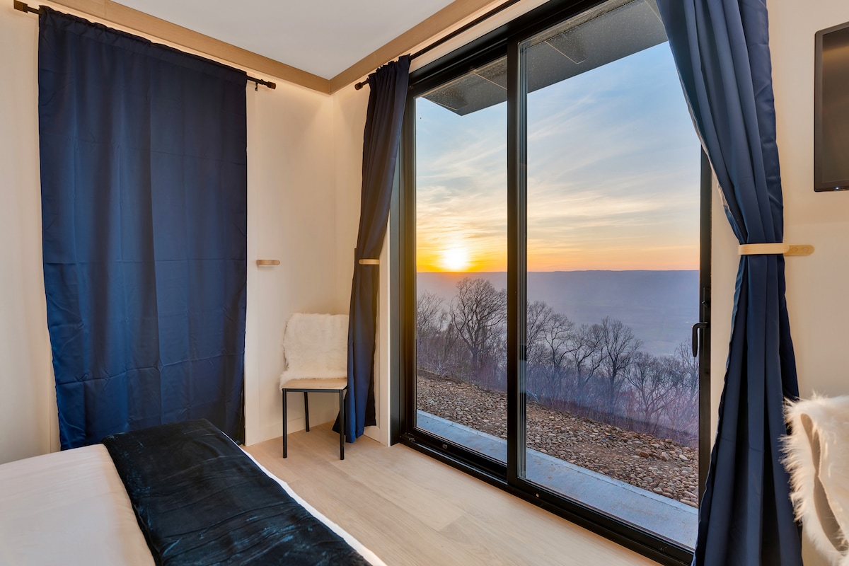 A serene bedroom is highlighted by large glass doors that open to a stunning sunset view. Soft light filters through dark curtains, while a minimalist chair stands by the window, inviting relaxation in the tranquil atmosphere.