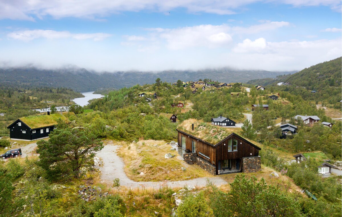 A panoramic view captures a charming holiday home set against a backdrop of rolling hills and a shimmering lake. The landscape is dotted with other cabins and greenery, offering a serene connection to nature under a cloudy sky.