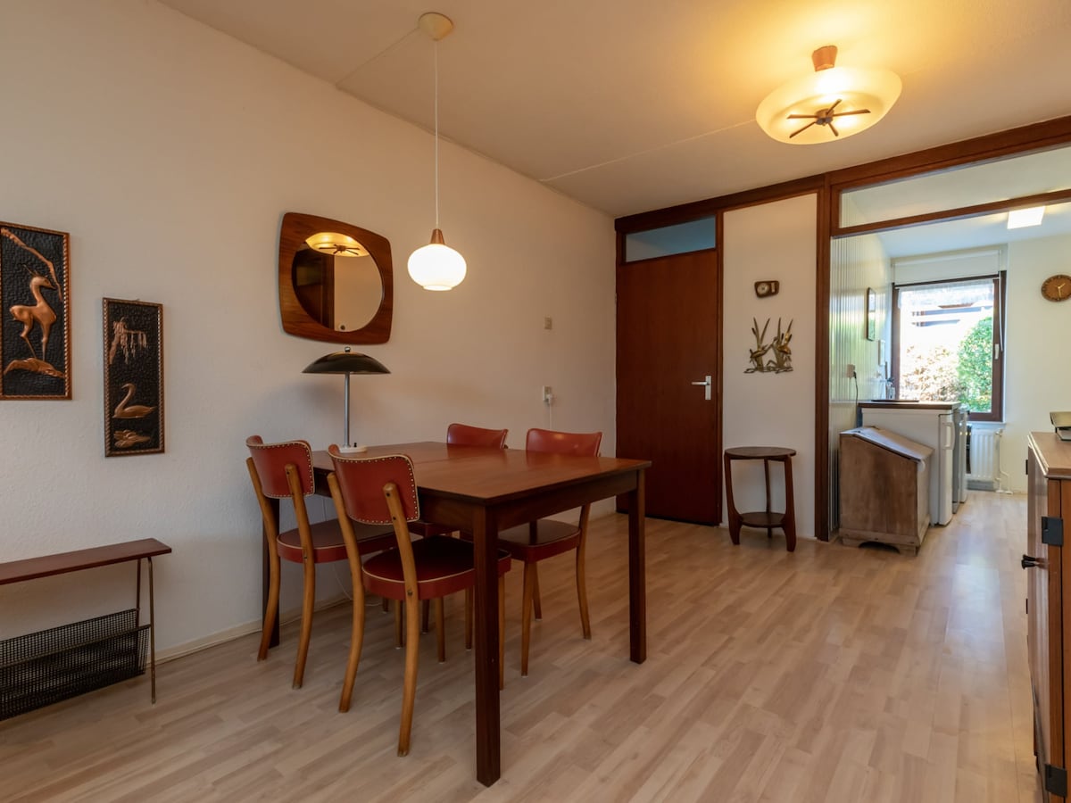 A dining area is presented with a wooden table surrounded by four red and natural wood chairs. A round ceiling light is visible above, with wall art displayed on either side. A doorway leads to a well-lit kitchen, framed by a window showing greenery outside.