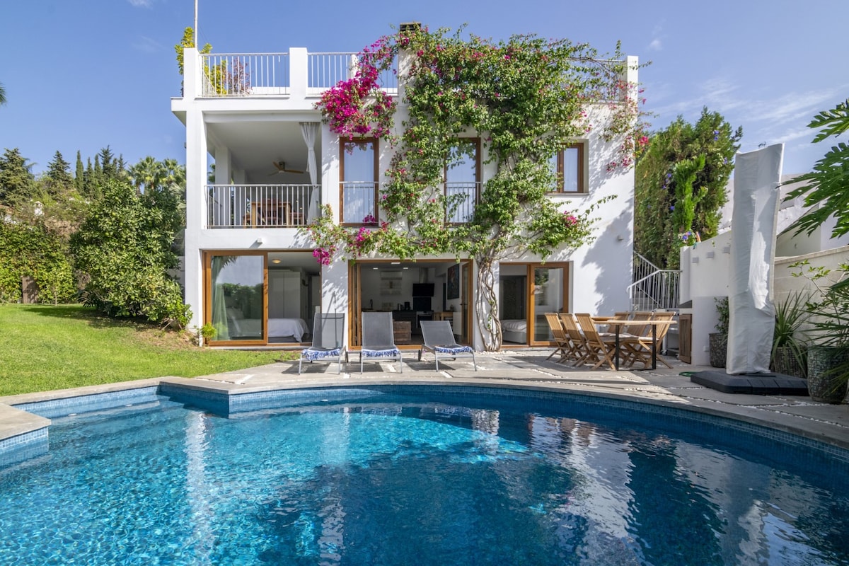 A spacious villa is set against a backdrop of vibrant greenery, featuring a sparkling swimming pool surrounded by lounge chairs. The two-story structure is adorned with flowering vines and offers large glass doors that provide access to an outdoor dining area, showcasing a relaxing outdoor environment.