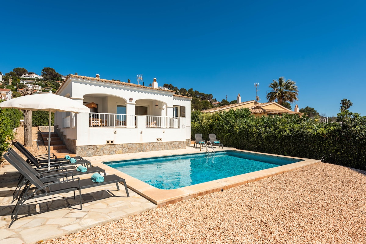 A private pool is surrounded by a landscaped area, featuring a spacious sun terrace with sunbeds and an umbrella. The house in the background is single-level, showcasing a light exterior and large windows that invite natural light.