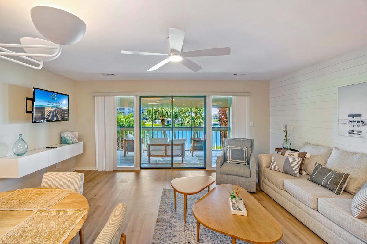 A light-filled living area features a comfortable sofa and an armchair, positioned near a coffee table. A dining set is visible, offering a relaxed space for meals. Floor-to-ceiling sliding glass doors invite views of lush greenery and provide access to the outdoors.
