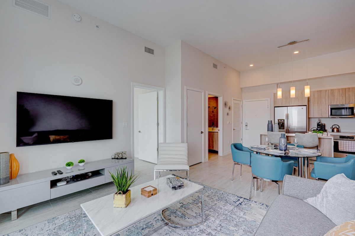 A modern living area showcases a large flat-screen television mounted on a light-colored wall. The space features a dining table with blue chairs, a sleek gray sofa, and a marble coffee table. A minimalist décor enhances the open layout, with views of connected rooms visible.