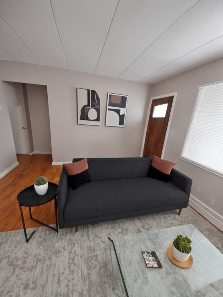 A modern living area features a sleek gray couch with decorative pillows, positioned near a glass coffee table. Art pieces adorn the wall above the couch, while natural light filters through the window. A simple side table with a potted plant adds a touch of greenery.