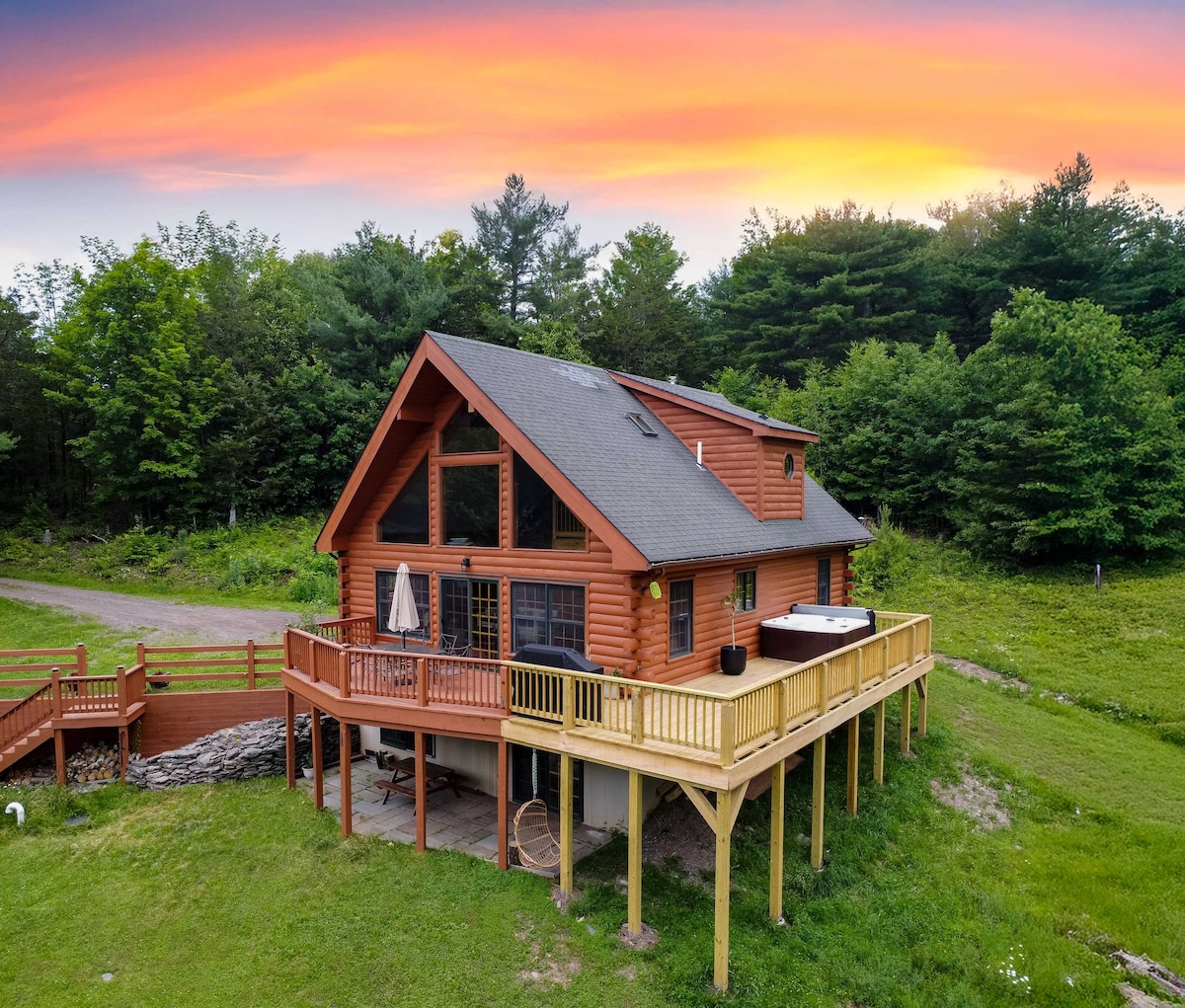 A two-story chalet features a wraparound deck with seating and an umbrella. Large windows provide views of the surrounding greenery, while a hot tub is positioned on the deck. The sunset casts a warm glow over the rustic log structure.