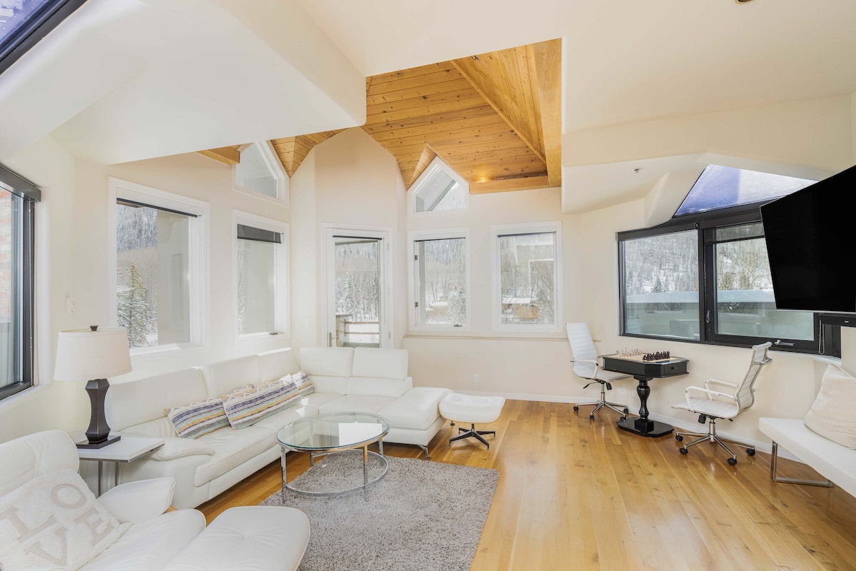 A spacious living area features large windows allowing natural light to fill the room. A modern white sectional sofa and a glass coffee table are positioned on a soft area rug. A sleek desk with a chair sits in a corner, and a flat-screen TV is mounted on the wall.