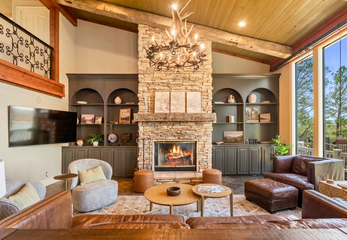 A spacious living area showcases a stone fireplace at its center, flanked by built-in shelves displaying decorative items. Comfortable seating is arranged around a coffee table, with large windows inviting natural light and offering views of the surrounding greenery.